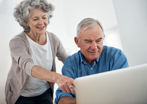 Vrouw van middelbare leeftijd en oudere man in gesprek aan tafel. Ze zijn bezig met een laptop. Mevrouw legt wat uit aan de meneer