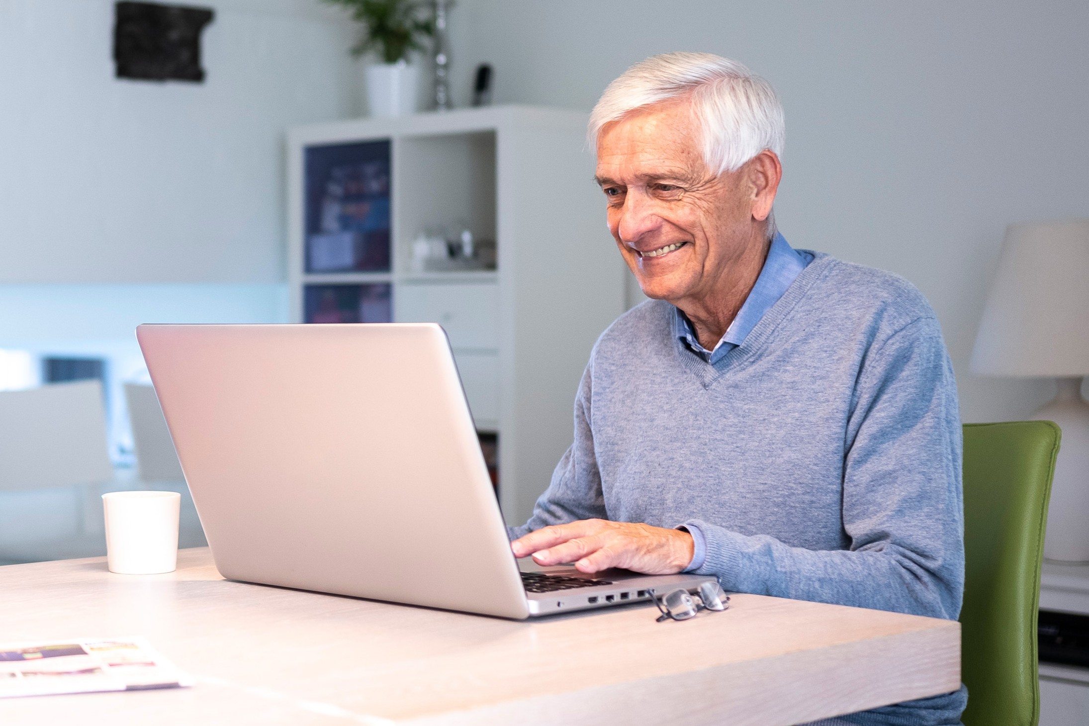 Blije senior man aan tafel in een kamer achter zijn laptop.