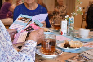 Oudere vrouwen zitten aan een tafel met thee en gebak, terwijl ze kleurrijke informatieve kaartjes bekijken over veilig bankieren.