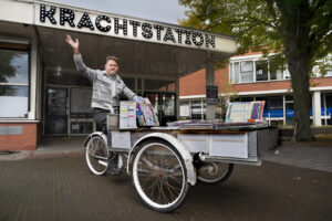 Coen Mobach op een bankfiets voor buurtcentrum "Krachtstation". Hij zwaait naar de fotograaf.