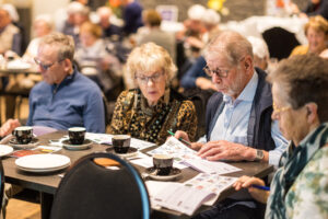 Groep senioren zit aan tafels tijdens een workshop over bankzaken, waar ze aandachtig materialen doornemen aan tafel met koffie.