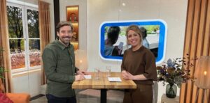Twee personen staan aan een hoge tafel in een warme studio‑setting, in gesprek met papieren en glazen water voor zich, met een groot scherm op de achtergrond.