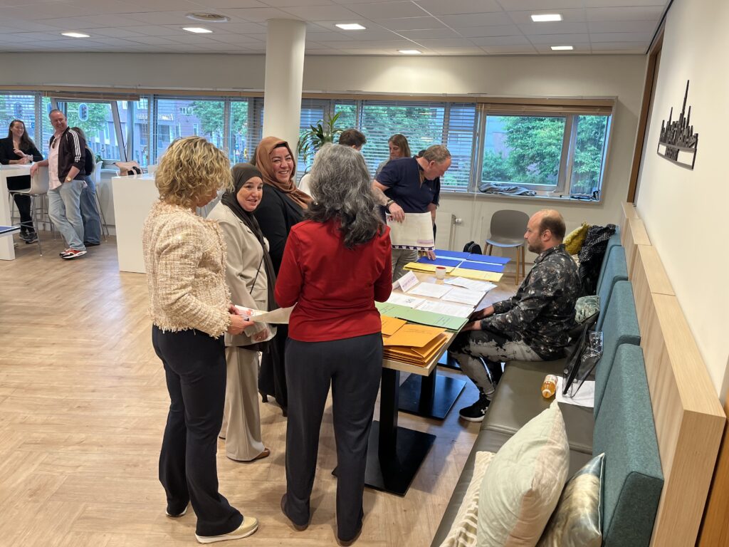 Groep personen bespreekt gekleurde kaarten rond een tafel tijdens een interactieve werksessie van het Verbindingsspel in een lichte kantoorruimte.