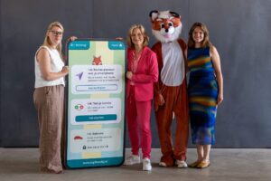 Vier personen poseren naast een groot smartphone-display van de DigiHandig-app. Van links naar rechts: Liesbeth Dewynn (Febelfi n), DigiVos (mascotte), Miriam van Osten (Toegankelijk Bankieren, namens de Nederlandse betaalsector) en Marit Huisman (DigiHandig). Op het scherm is een DigiHandig-les over routes plannen met een smartphone te zien.