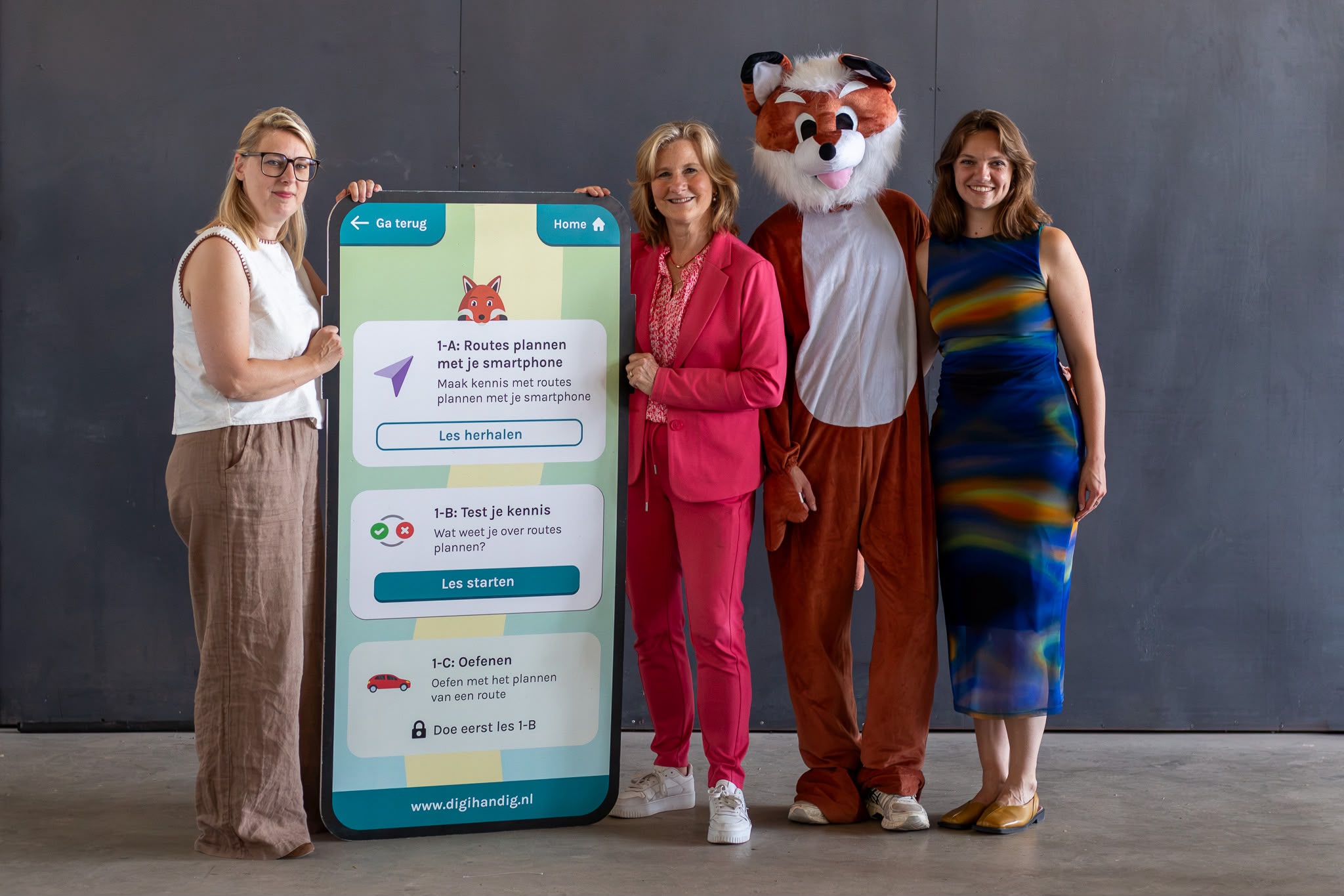 Vier personen poseren naast een groot smartphone-display van de DigiHandig-app. Van links naar rechts: Liesbeth Dewynn (Febelfi n), DigiVos (mascotte), Miriam van Osten (Toegankelijk Bankieren, namens de Nederlandse betaalsector) en Marit Huisman (DigiHandig). Op het scherm is een DigiHandig-les over routes plannen met een smartphone te zien.