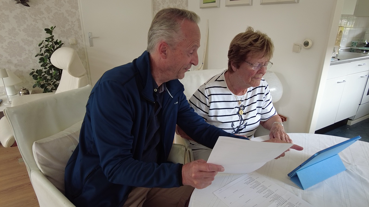 Man helpt oudere vrouw aan de keukentafel met haar bankzaken, onder andere met papieren .